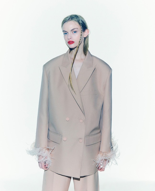 Fashion portrait of a woman in a beige blazer with feathered cuffs and matching pants, posing against a clean white backdrop.