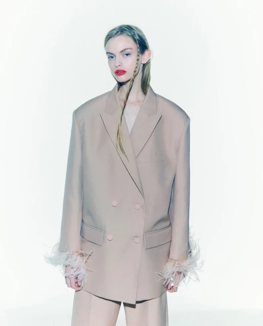 Fashion portrait of a woman in a beige blazer with feathered cuffs and matching pants, posing against a clean white backdrop.