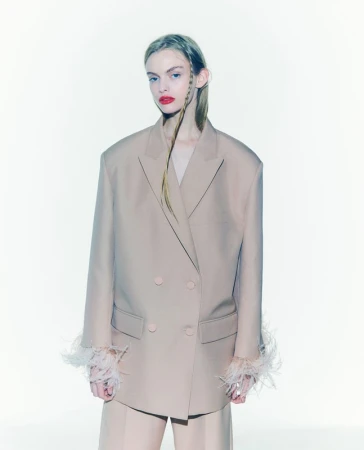 Fashion portrait of a woman in a beige blazer with feathered cuffs and matching pants, posing against a clean white backdrop.