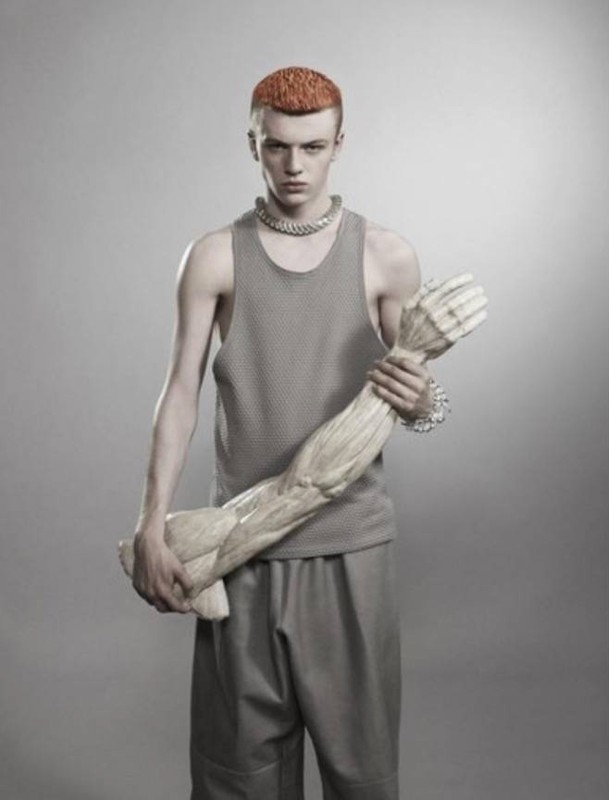 A young adult male poses in a studio, wearing a gray tank top and pants, holding a prop of a human arm, with a gray background.