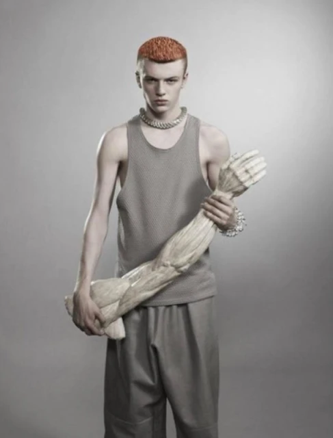 A young adult male poses in a studio, wearing a gray tank top and pants, holding a prop of a human arm, with a gray background.
