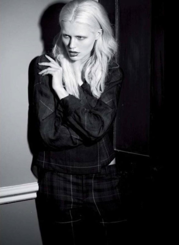 A fashion shot of a woman with platinum hair, wearing a patterned blouse and pants, stands against a wall in a dimly lit indoor setting.