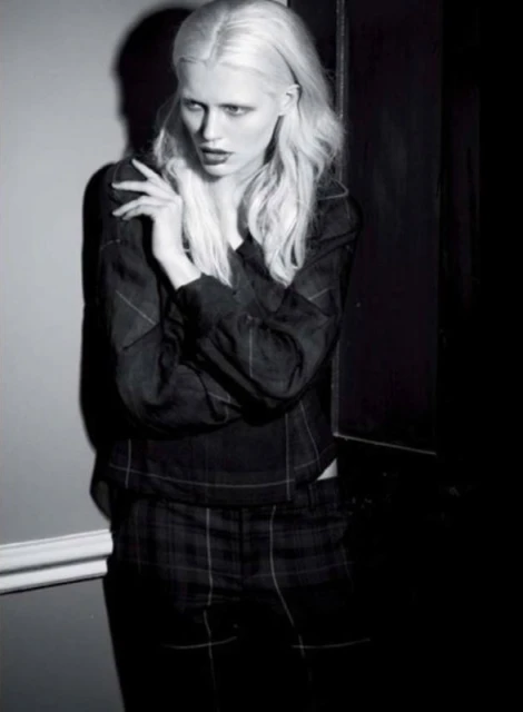 A fashion shot of a woman with platinum hair, wearing a patterned blouse and pants, stands against a wall in a dimly lit indoor setting.