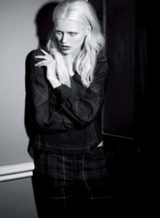 A fashion shot of a woman with platinum hair, wearing a patterned blouse and pants, stands against a wall in a dimly lit indoor setting.