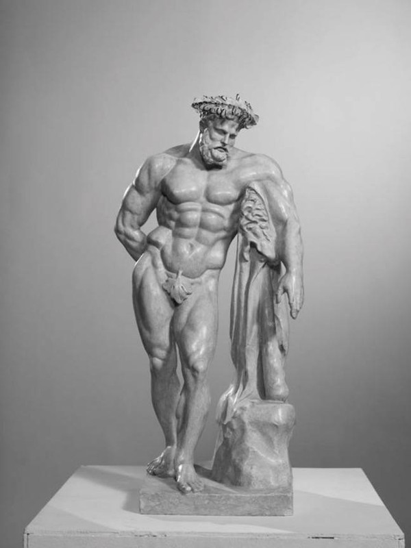 Monochrome statue of a muscular man with a beard, wearing a wreath, draped cloth, and standing on a podium in a neutral setting.