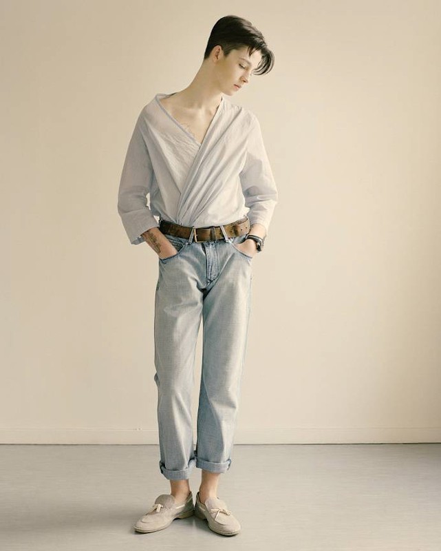 Full-body shot of a young adult posing indoors wearing a light blue collared shirt, jeans, belt, and loafers against a neutral backdrop.
