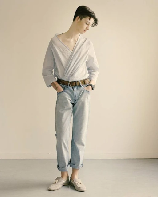 Full-body shot of a young adult posing indoors wearing a light blue collared shirt, jeans, belt, and loafers against a neutral backdrop.