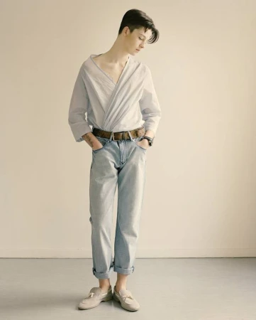 Full-body shot of a young adult posing indoors wearing a light blue collared shirt, jeans, belt, and loafers against a neutral backdrop.