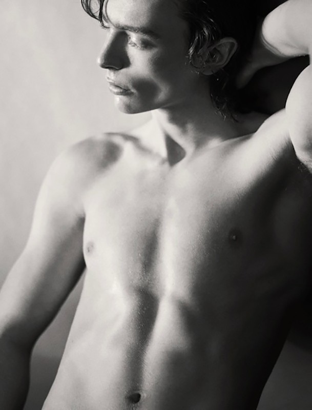 Portrait of a shirtless young man in profile with dark wavy hair and one hand raised behind his head.