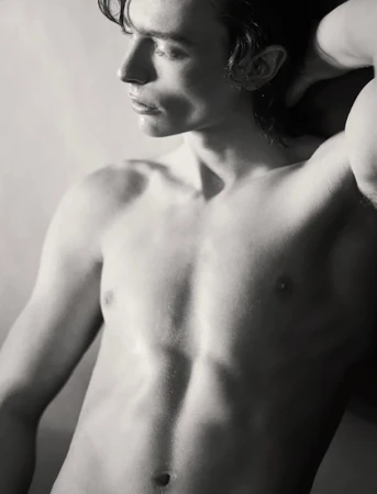 Portrait of a shirtless young man in profile with dark wavy hair and one hand raised behind his head.