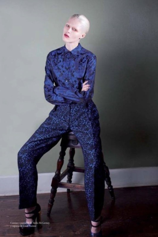 Model with platinum blonde hair sitting on a wooden stool wearing a patterned blue button-up shirt and matching trousers.