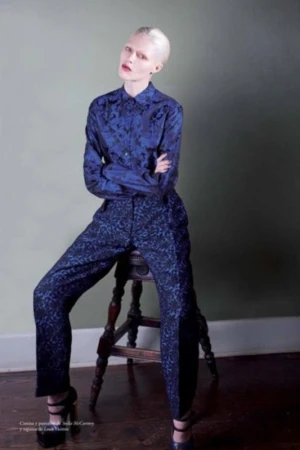Model with platinum blonde hair sitting on a wooden stool wearing a patterned blue button-up shirt and matching trousers.