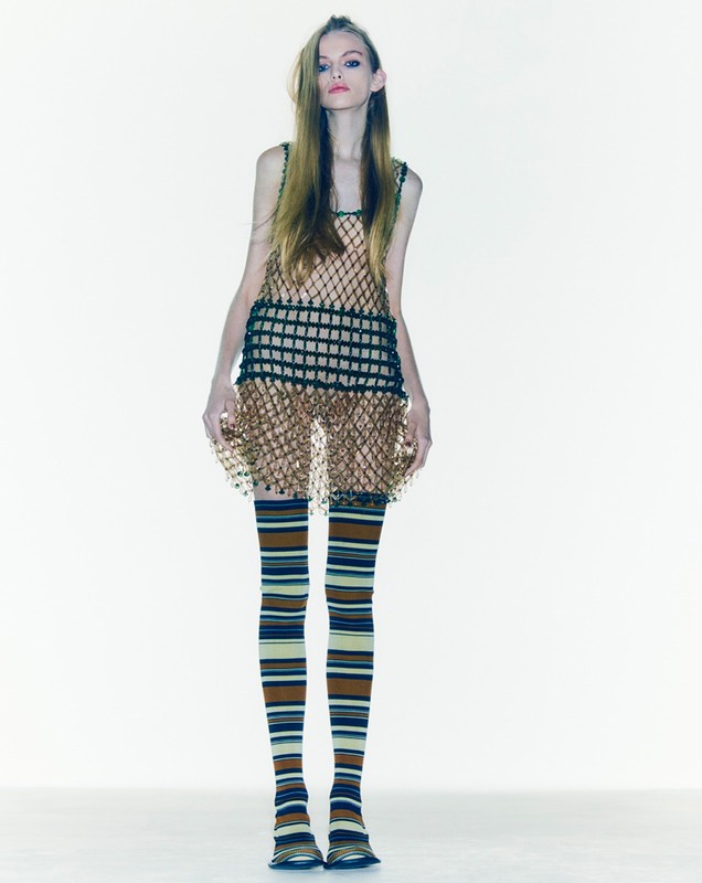 A slender fashion model in a net dress poses in a studio setting, wearing striped tights and flats, presented against a white backdrop.