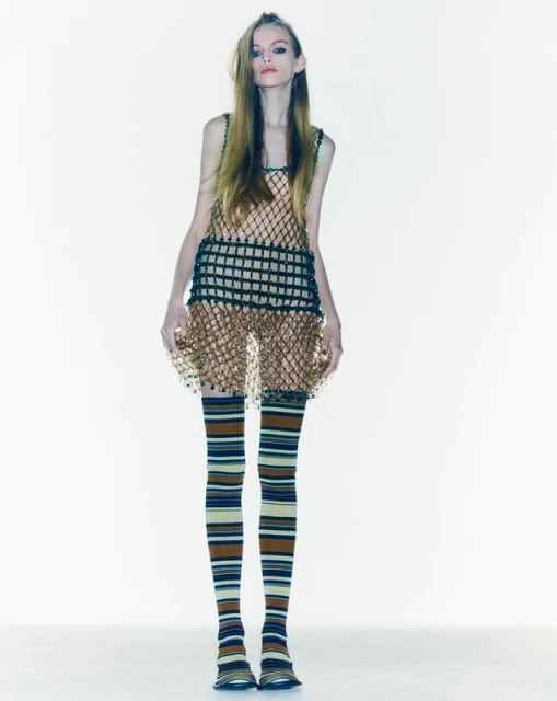 A slender fashion model in a net dress poses in a studio setting, wearing striped tights and flats, presented against a white backdrop.