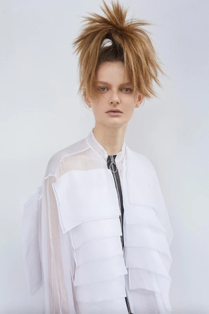 Young adult woman with updo hairstyle poses in semi-sheer white jacket against white backdrop in portrait-style studio shot.