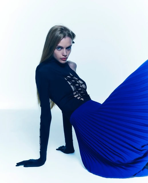 A fair-skinned woman in a black long-sleeved top and royal blue pleated skirt is posing against a white backdrop in a studio setting.