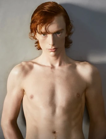 Close-up portrait of a shirtless young man with red hair looking directly forward against a grey studio backdrop