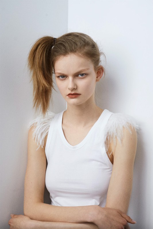Portrait of a young adult female in a white top with feathered shoulder accents posing against a plain white corner background.