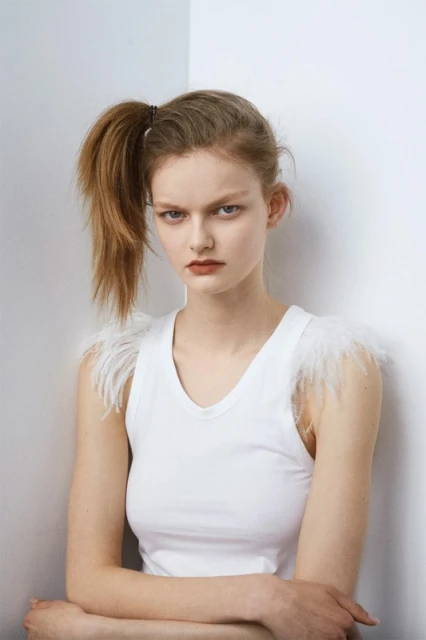 Portrait of a young adult female in a white top with feathered shoulder accents posing against a plain white corner background.