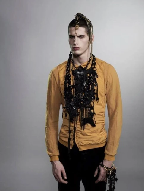 Portrait of a young adult male with dark hair, wearing an elaborate black beaded necklace and a yellow jacket against a gray background.