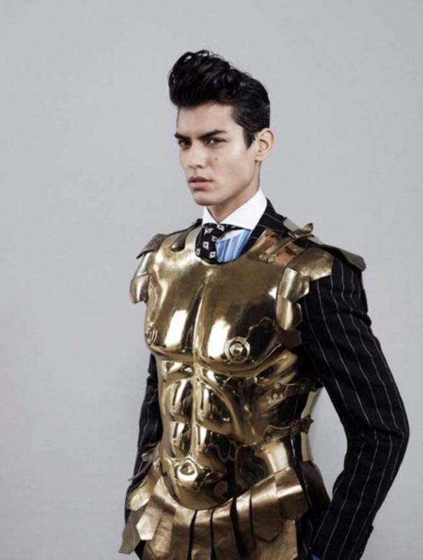 Male model poses in a formal pinstripe suit with a gold-colored, Roman-style breastplate in a studio with a neutral backdrop.
