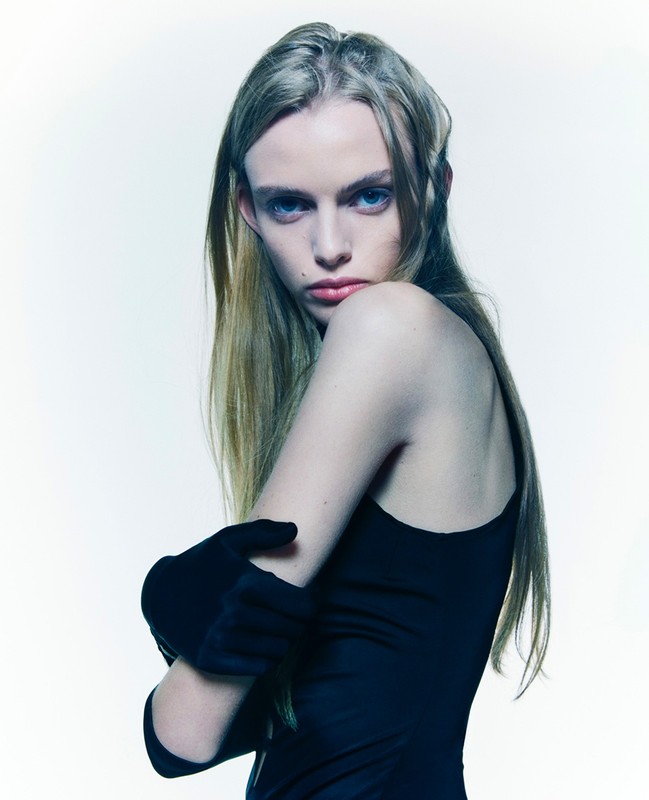 Indoor portrait of a blonde adult female fashion model with her arms crossed, wearing black gloves and a dark dress on a white background.