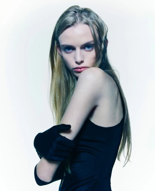 Indoor portrait of a blonde adult female fashion model with her arms crossed, wearing black gloves and a dark dress on a white background.