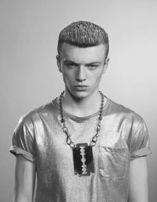 A young adult male gazes intensely into the camera, sporting a buzzcut and an edgy razorblade necklace, all set against a neutral backdrop.