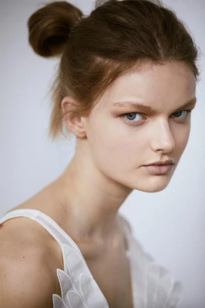Close-up portrait of a woman with hair styled in a top knot wearing a white sleeveless dress against a white background.