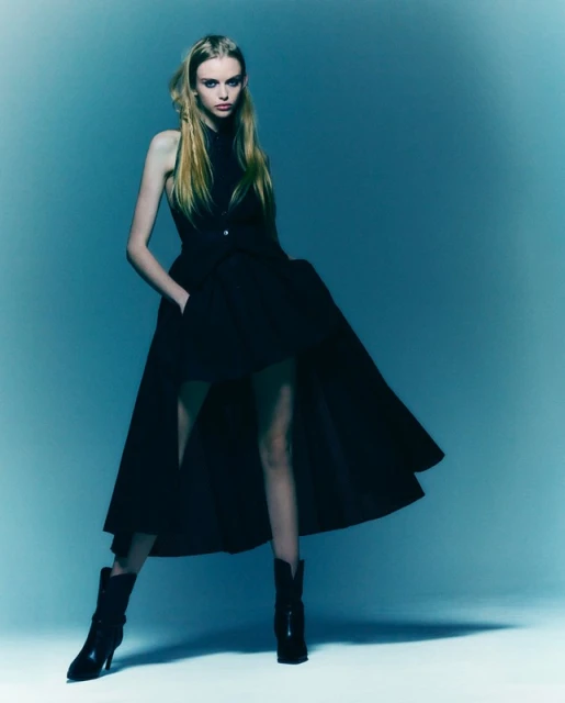 Full-length portrait of a woman in a black dress and boots posing against a cyan background in a studio fashion shot.