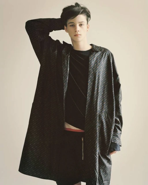Portrait of a young adult male model with dark hair wearing a patterned black coat and shorts in a photo studio setting, looking at the camera.