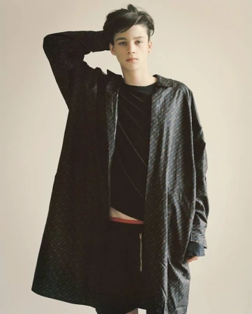 Portrait of a young adult male model with dark hair wearing a patterned black coat and shorts in a photo studio setting, looking at the camera.