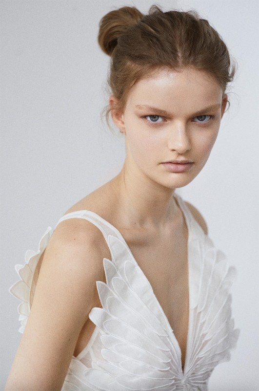 Close-up portrait of a woman with fair skin and her hair in a bun, wearing a white dress with floral detailing against a white background.