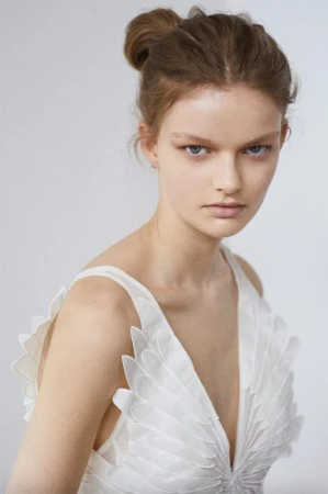 Model with hair styled in a high bun wearing a white v-neck dress with petal appliques against a seamless background