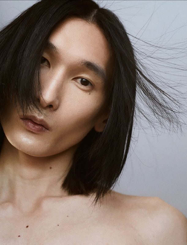 Close-up portrait of a man with long dark hair blowing across his face against a neutral grey background