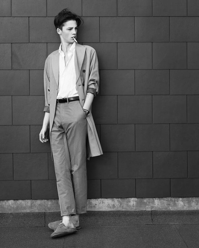 Black and white portrait of a young man in a coat smoking a cigarette, posing against a dark tiled wall in an urban outdoor setting.