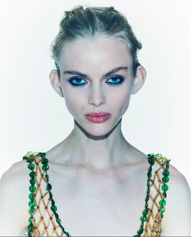 Close-up portrait of a young adult woman with a beaded top against a white background, emphasizing her makeup and jewelry.