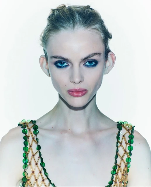 Close-up portrait of a young adult woman with a beaded top against a white background, emphasizing her makeup and jewelry.