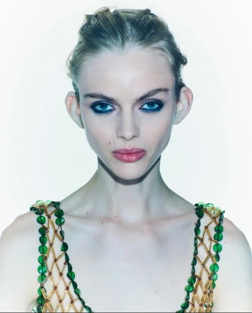 Close-up portrait of a young adult woman with a beaded top against a white background, emphasizing her makeup and jewelry.