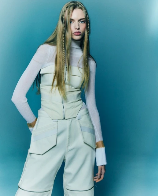 Fashion portrait of a young woman in a pale yellow top and pants with braids in her long blonde hair against a blue backdrop.