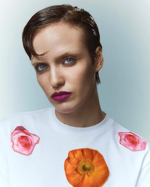 Close-up portrait of a woman with short, slicked-back hair, wearing a white t-shirt with flower appliques against a gradient background.
