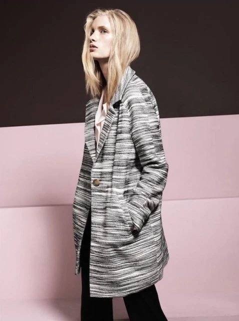 A fair-skinned woman with blonde hair wears a striped grey overcoat and black slacks against a layered pink and brown backdrop.