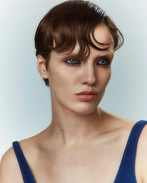 Close-up portrait of a young adult woman with dark, waved hair and blue eye makeup wearing a blue tank top against a light background.