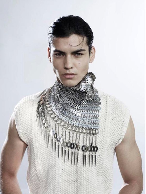Close-up of a male model with dark hair wearing a sleeveless textured top and a unique silver metal bib necklace in a studio setting.