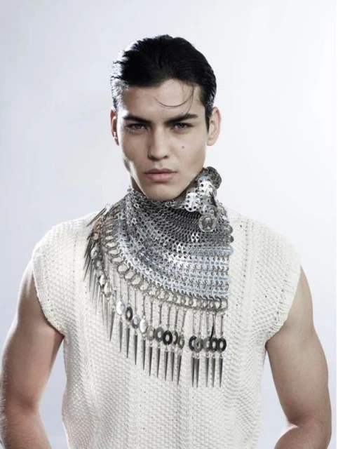 Close-up of a male model with dark hair wearing a sleeveless textured top and a unique silver metal bib necklace in a studio setting.