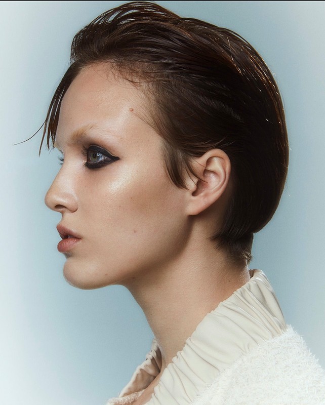 Close-up profile of a woman with short dark hair, heavy eyeliner, pale skin, and a neutral expression against a light blue background.