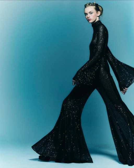 A fashionable adult female model poses in a black, sequined jumpsuit with bell sleeves and pants in front of a graduated blue background.