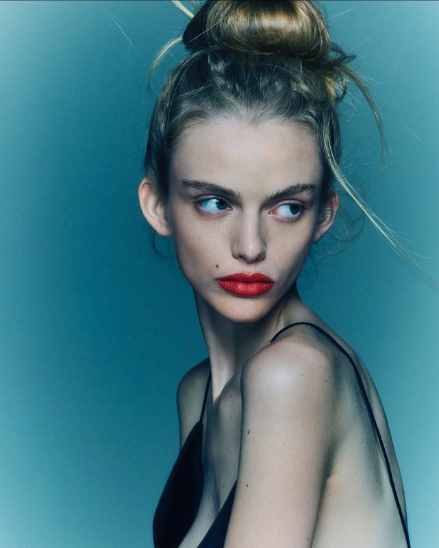 Close-up portrait of a young woman with blonde hair in a bun, wearing red lipstick and a black tank top against a teal background.