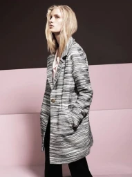 Model with blonde hair wearing a patterned grey coat and dark trousers against a pink and dark brown studio wall.