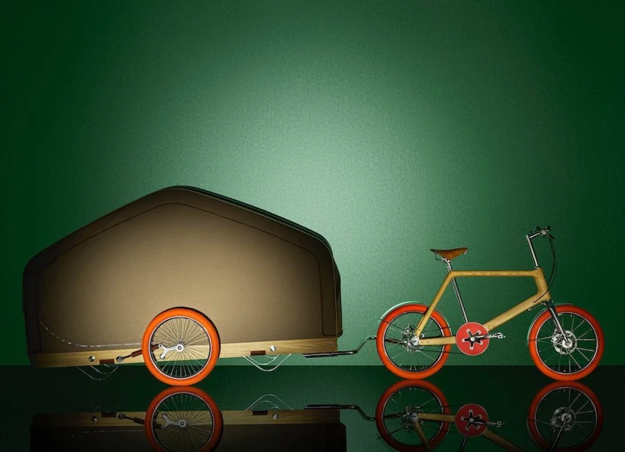 A bicycle and trailer, likely for transporting goods, sit on a reflective surface against a green backdrop in a studio setting.